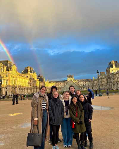 A group of soggy students dress in warm, casual attire as they stand in front of the Louvre on a rainy day. A double rainbow appears in the sky above their heads.