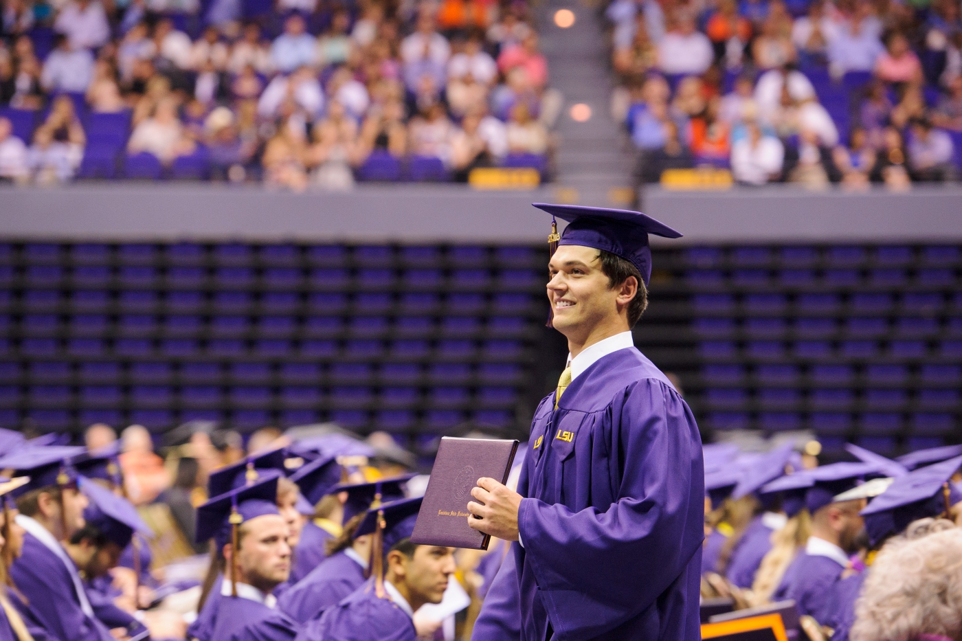 Student in graduating purple cap and gown stands and smiles with diploma among seated students at ceremony. 