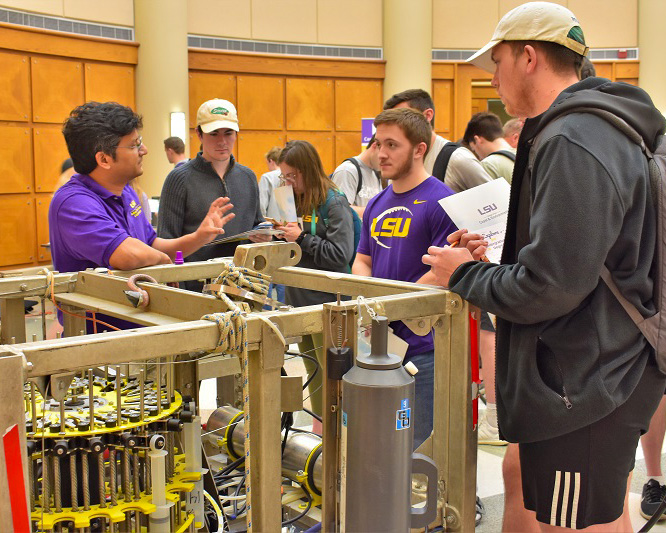 a man stands next to a large piece of scientific equipment talking to two students