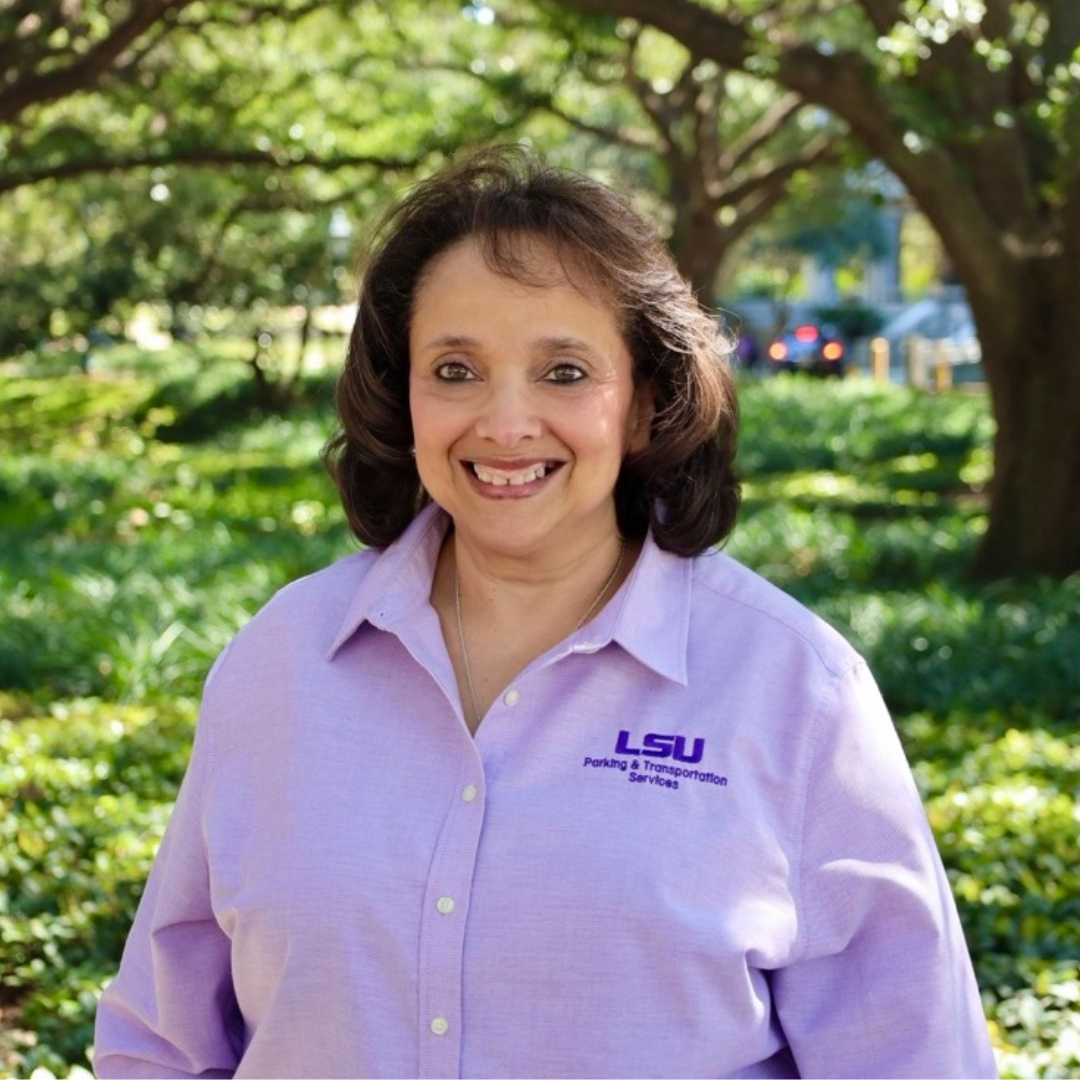 Woman in purple shirt smiling and standing in front of a tree and bushes